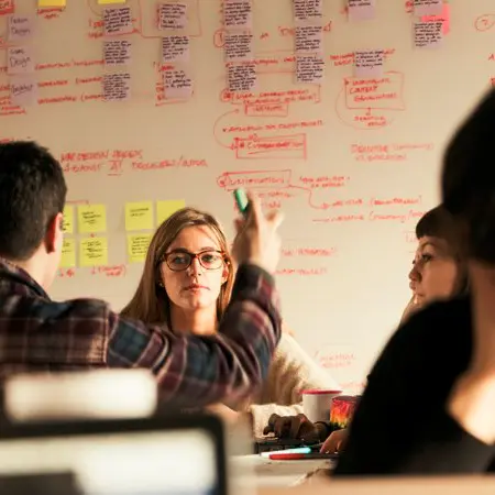 The image shows students sitting at a desk in front of a white board.